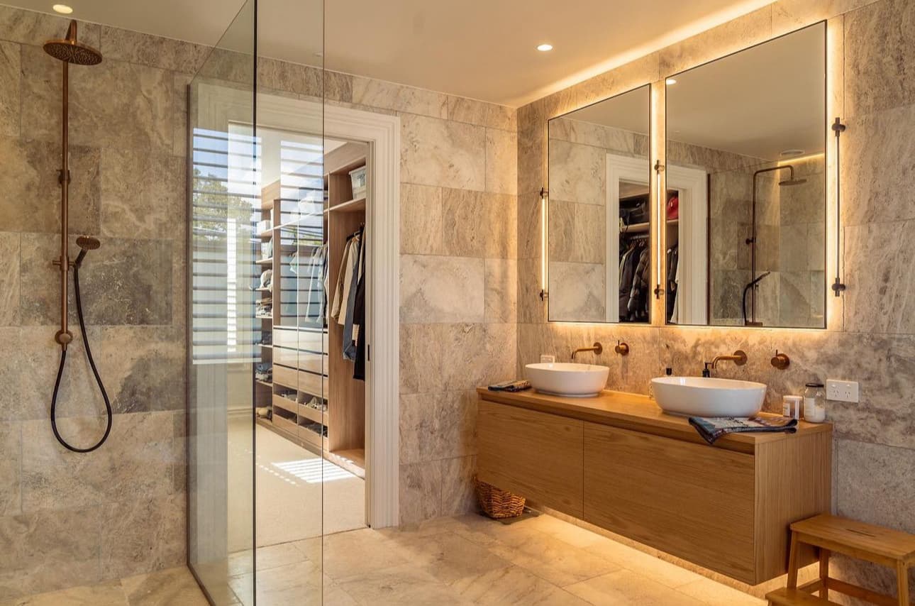 Luxury bathroom with beige natural stone tiles, glass shower, backlit mirrors, double vanity, and walk-in closet