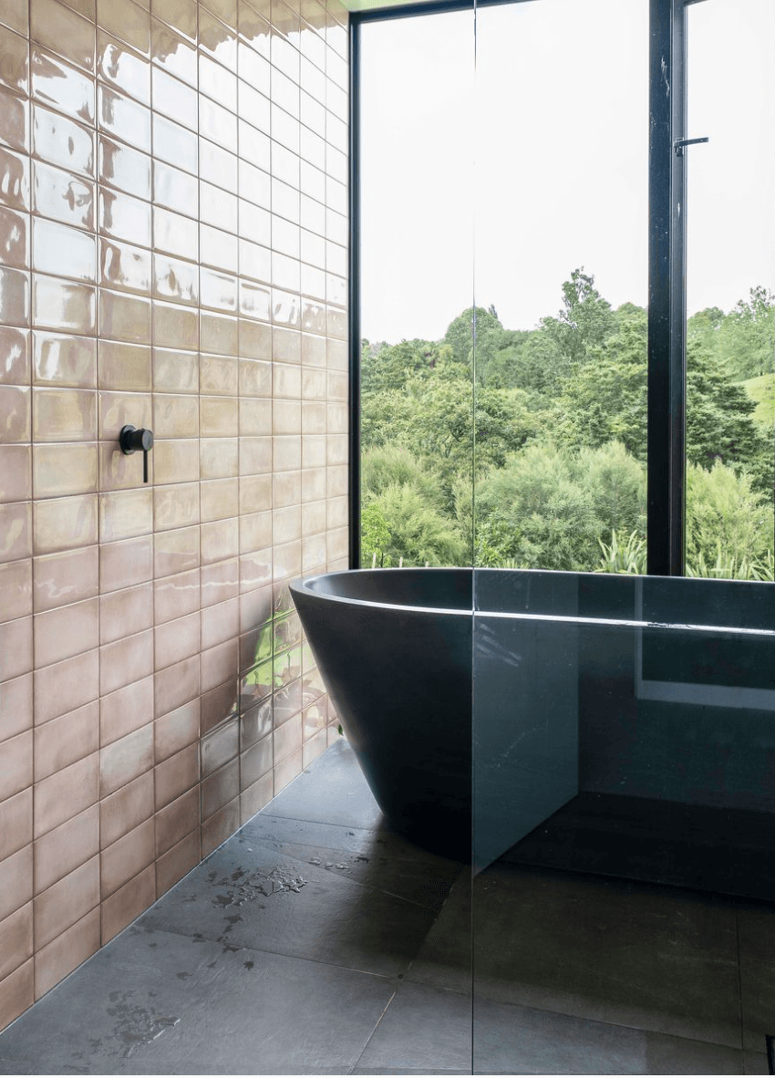 Dramatic bathroom with glossy blush subway tiles, black freestanding bathtub, and floor-to-ceiling windows overlooking native bush