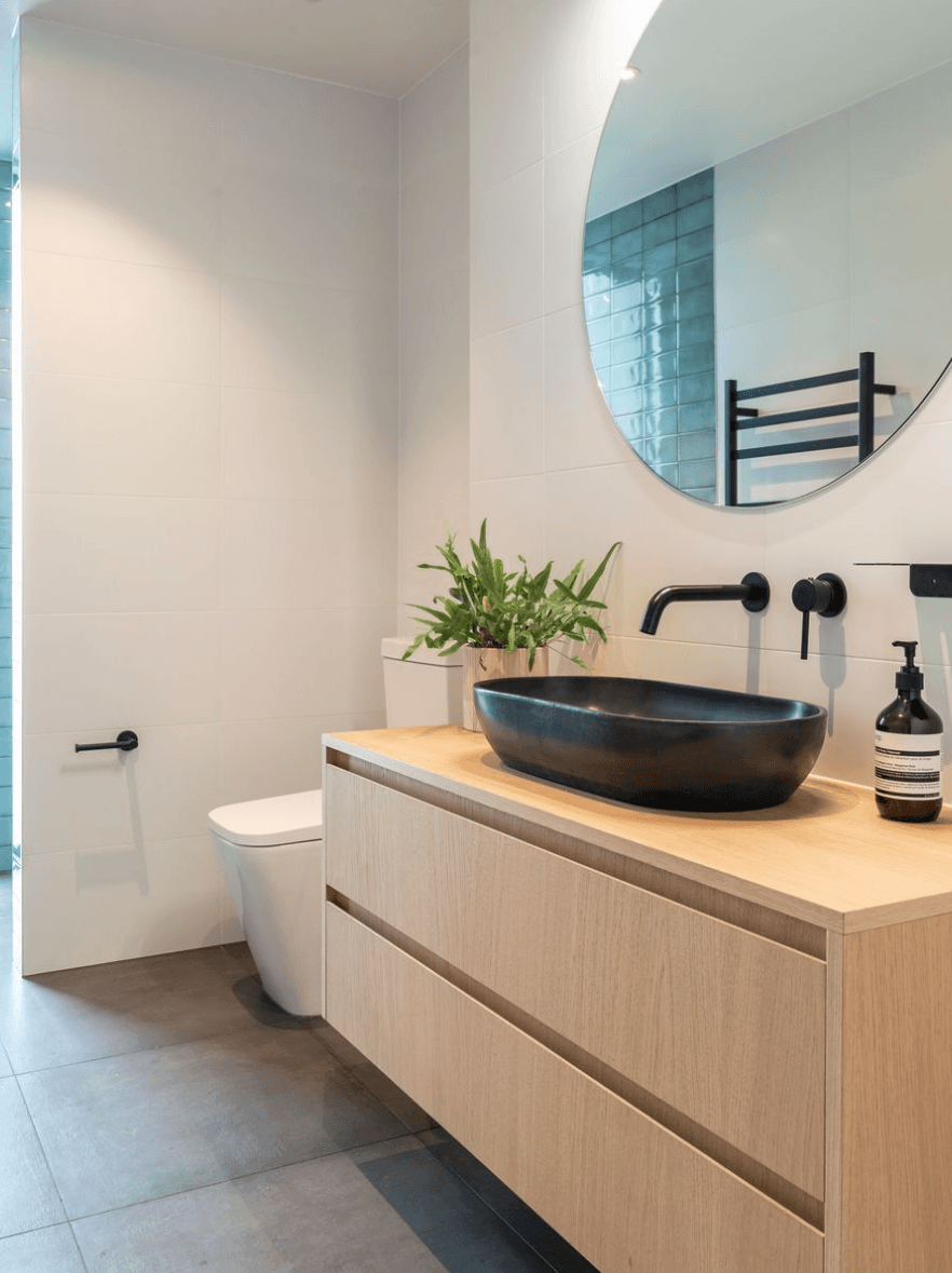Clean modern bathroom with white wall tiles, wood-grain floating vanity, round mirror, and black vessel sink