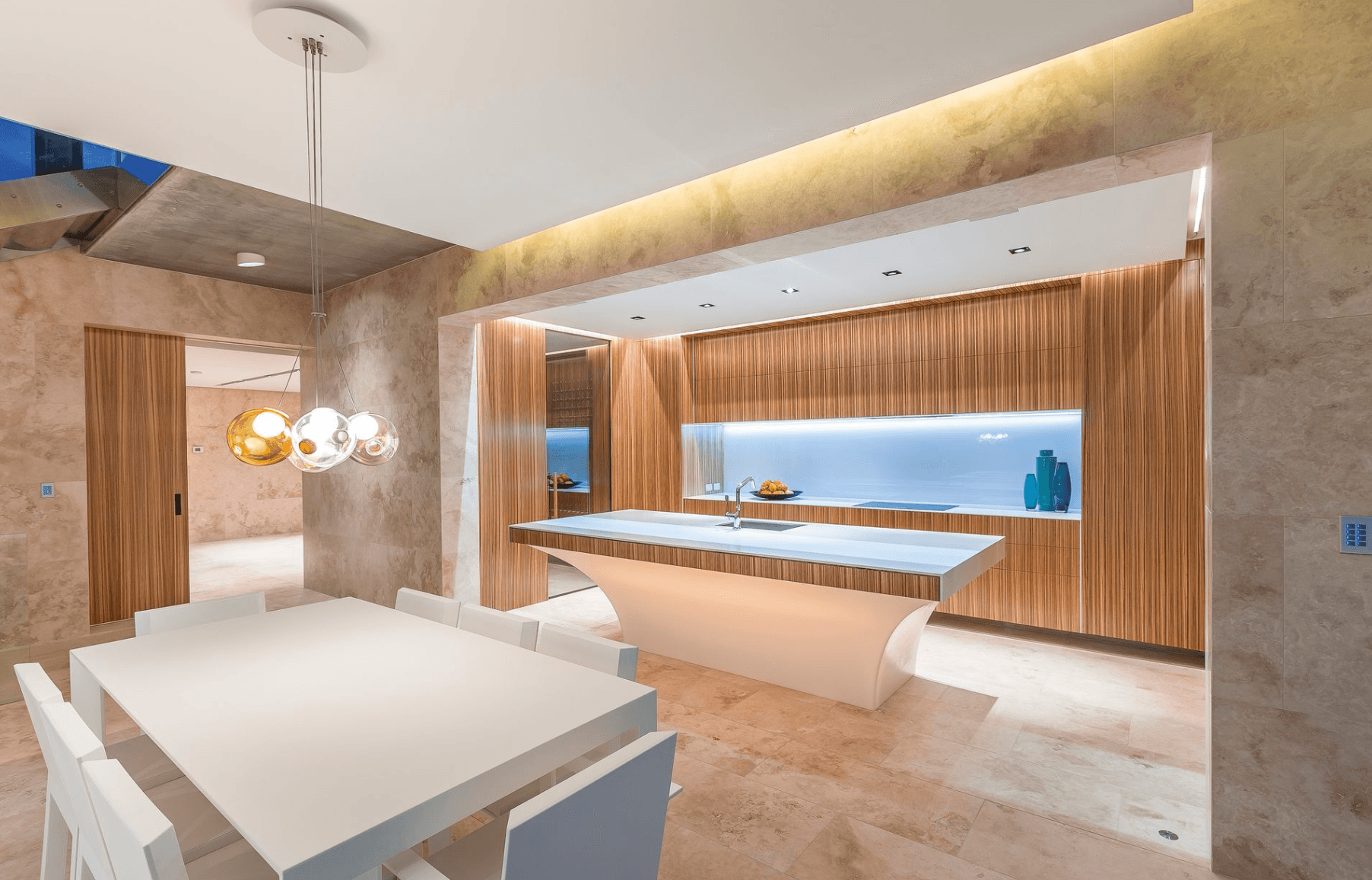 Luxury kitchen with natural stone walls and floors, wood-paneled cabinetry, sculptural curved white island, and colored-glass cluster pendant over a white dining table